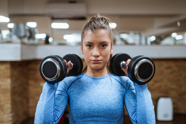 Μια νεαρή όμορφη γυναίκα σε μπλε αθλητικές ασκήσεις εξοπλισμού στο γυμναστήριο με βάρη - Φωτογραφία, εικόνα