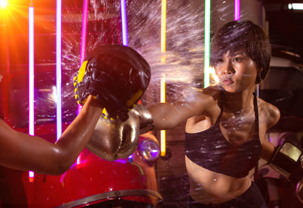 Όμορφη εικοσάρα ασιατική γυναίκα τρένα punching με τον προπονητή στο Silver γάντια μόδας Mitts. Office Girl άσκηση στο σύγχρονο πολύχρωμο Neon Muay Thai Πυγμαχία Γυμναστήριο φόντο με νερό ιδρώτα splash φως - Φωτογραφία, εικόνα