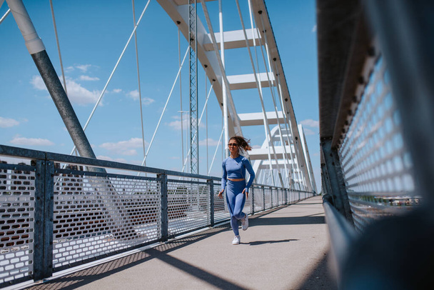 A young beautiful caucasian girl in blue sports equipment is running on the bridge. Training in nature - 写真・画像