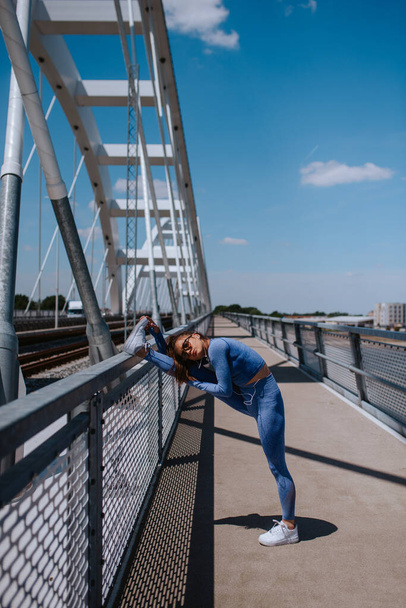 Ein junges hübsches kaukasisches Mädchen in blauer Sportausrüstung streckt ihr Bein auf der Brücke aus. Ausbildung und gesundes Leben - Foto, Bild