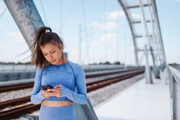 Πορτρέτο μιας νεαρής όμορφης καυκάσιας κοπέλας με μπλε αθλητικό εξοπλισμό με ένα τηλέφωνο στο χέρι στη γέφυρα - Φωτογραφία, εικόνα