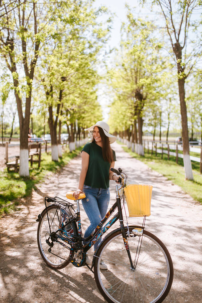  Μια όμορφη νεαρή γυναίκα με ψάθινο καπέλο, ποδήλατο και λουλούδια στέκεται στο πάρκο. Η ζωή σε ένα χωριό - Φωτογραφία, εικόνα