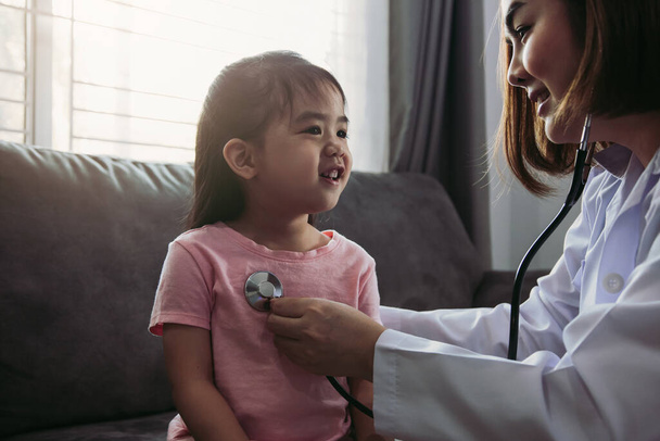 Selbstbewusste asiatische Gesundheitsexpertin besucht ihre Patientin mit einem Stethoskop, während eine junge Patientin zu Hause gut untersucht wird. - Foto, Bild