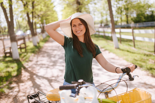 A beautiful young woman with a straw hat, bicycle and flowers is standing in the park. Life in a village - Фото, зображення