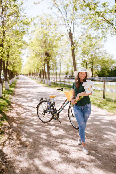 Μια όμορφη νεαρή γυναίκα με ψάθινο καπέλο, ποδήλατο και λουλούδια στέκεται στο πάρκο. Η ζωή σε ένα χωριό - Φωτογραφία, εικόνα