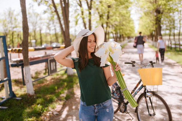 Μια όμορφη νεαρή γυναίκα με ψάθινο καπέλο, ποδήλατο και λουλούδια στέκεται στο πάρκο. Η ζωή σε ένα χωριό - Φωτογραφία, εικόνα