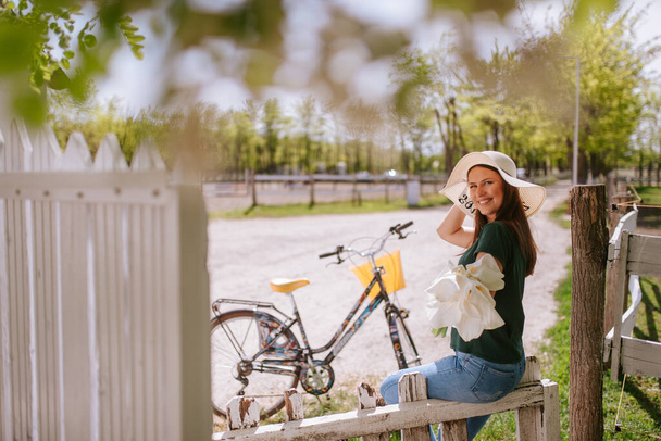 Een jonge mooie blanke vrouw met een strohoed, een boeket calla en een fiets naast een hek in het dorp - Foto, afbeelding