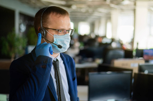 A man in latex gloves and a medical mask is talking to a client over the headset. A male office manager answers calls. Work during the coronavirus. White-collar tie and jacket. - Fotó, kép