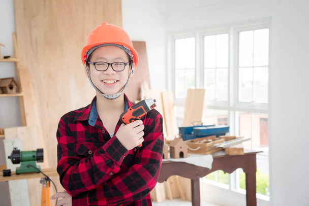 młody azjatycki chłopiec noszący kask i trzymający stolarz narzędzie z uśmiechem stojący w stolarni. - Zdjęcie, obraz