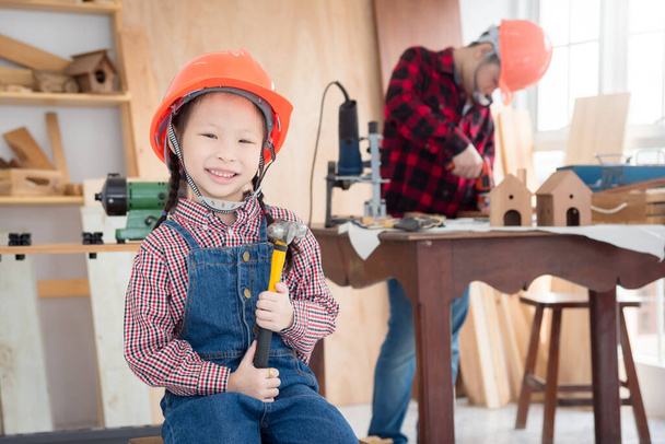 Küçük Asyalı kız miğfer takıyor ve elinde tebessümle çekiç tutuyor marangozhanede oturuyor.. - Fotoğraf, Görsel