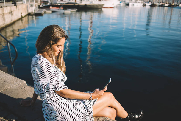 Attractive female tourist taking rest in port near mediterranean sea and using smartphone gadget for searching information and advices for travellers, Caucasian hipster girl reading received sms - Fotó, kép