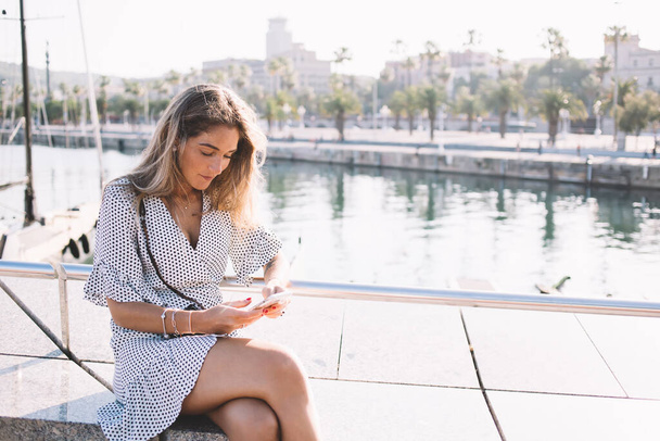 Attractive woman dressed in trendy sundress taking rest in Barcelona harbor using smartphone application for installing offline travel guide, beautiful female tourist sending sms message via phone - Zdjęcie, obraz