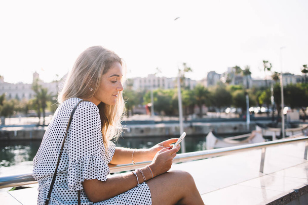 Beautiful millennial tourist writing text for publication in social networks connected to 4g wireless internet on modern mobile phone, attractive woman using smartphone gadget on leisure in port - Photo, Image