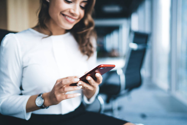 Crop blurred smiling modern business lady in formal outfit and wristwatch sitting and browsing mobile on blue blurry workspace background  - Фото, изображение