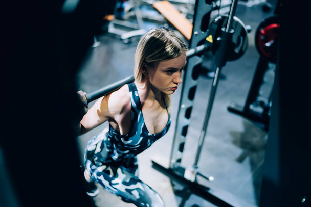 From above beautiful determined blonde fit female bodybuilder in sportswear and sneakers doing lunges with barbell while working out in gym  - 写真・画像