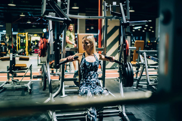 Determined fit female bodybuilder in tight urban camouflage sportswear and sneakers lifting heavy weights with chest lifting machine in gym - Φωτογραφία, εικόνα