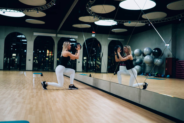 Side view of sporty female in black top and white leggings doing lunges in front of mirror with medicine ball in gym looking at own reflection  - Photo, Image