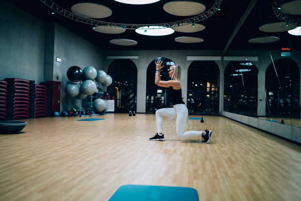 Side view of healthy fit female in black top and white leggings doing lunges with medicine ball during workout in gym - Zdjęcie, obraz