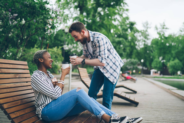 Side view of multiracial happy carefree couple cheering with coffee takeaway and joking while spending time in summer city park - Foto, Imagen