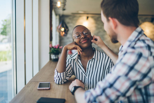 Side view of young man affectionately rubbing cheek of pleasant smiling African American girl friend in glasses at table in bright cafe - Photo, image