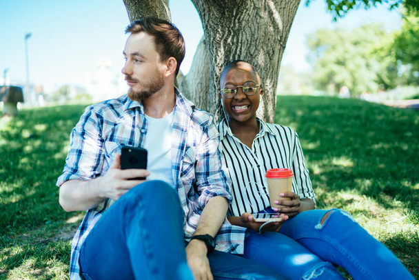 Calm male looking away inspired using mobile phone sitting under tree with African American female lover drinking coffee and listening to music in headphones cheerfully smiling - 写真・画像