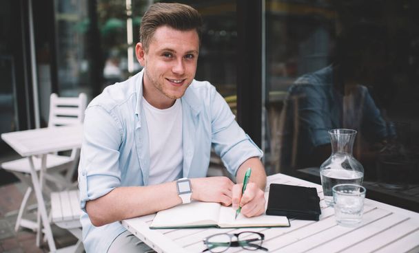 Young smiling male wearing blue shirt white t shirt and wrist watches making notes in notebook sitting at table with glasses and jug in outdoor terrace - Photo, image