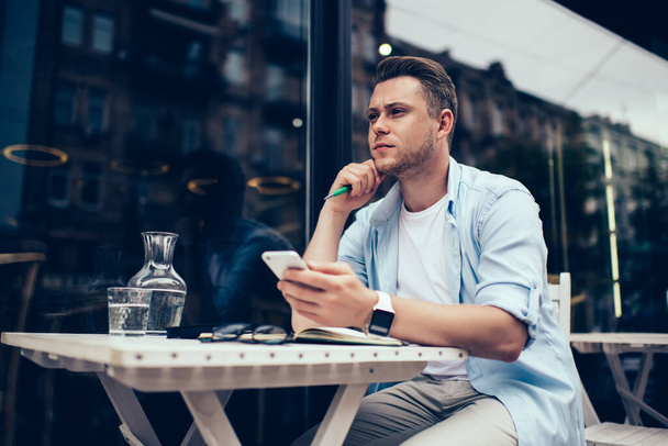 Concentrated intellectual male wearing blue shirt white t shirt and wrist watches texting on mobile phone sitting at table in outdoor terrac - Photo, Image