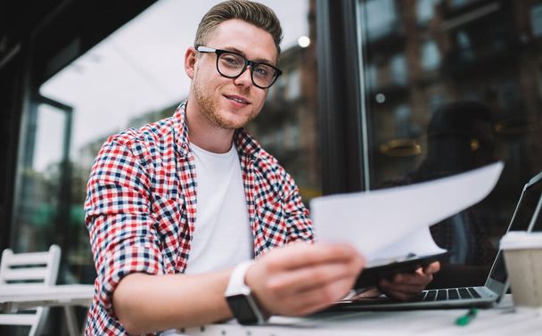 Cheerful freelancer in casual clothes and glasses exploring documents creating project sitting at table in street cafe looking at camera - Фото, зображення