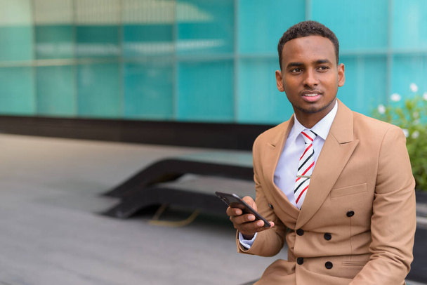 Счастливый молодой африканский бизнесмен с телефоном думает и сидит в городе на открытом воздухе
 - Фото, изображение