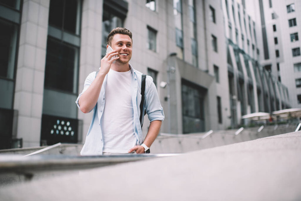 Low angle of young smiling stylish male in casual clothes having conversation on mobile phone while standing on stair in street and looking away - Φωτογραφία, εικόνα