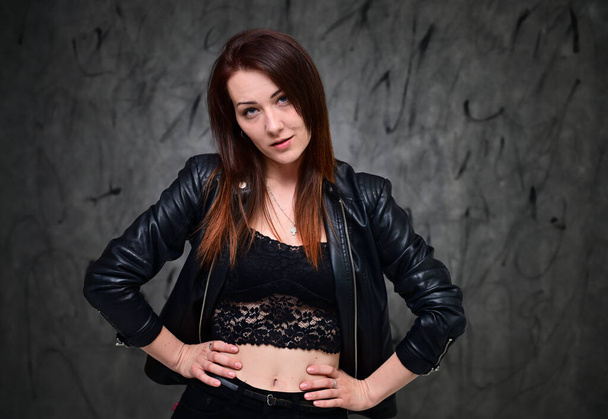 Studio portrait of a caucasian brunette girl with minimal makeup. A pretty model is standing on a gray background in a black jacket. - Photo, image