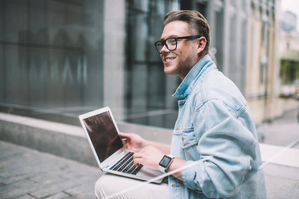 Side view of young stylish smiling spectacled male in smart watch wearing denim jacket typing on laptop while sitting in street and looking away - Photo, image