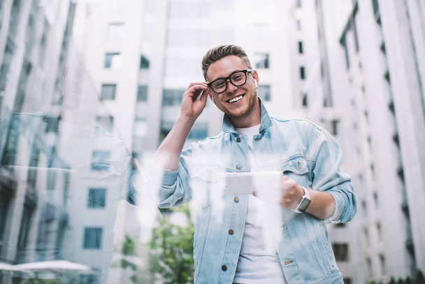 View from below of cheerful laughing man in casual clothes adjusting glasses talking to friend using video chat on tablet in street looking at camera - 写真・画像