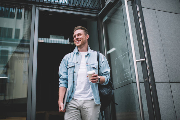 Content smiling young man in casual clothes with backpack taking cup of hot drink going out of coffee bar looking away - Foto, immagini