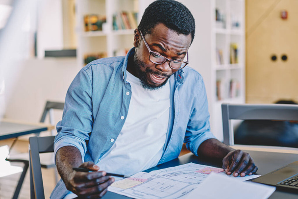 Astonished black guy in casual outfit and glasses checking draft while sitting at desk on blurred background of contemporary workplace - Zdjęcie, obraz