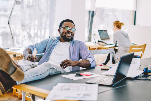 Cheerful African American guy with smartphone smiling and looking away while keeping legs on table and resting during break in office - Zdjęcie, obraz