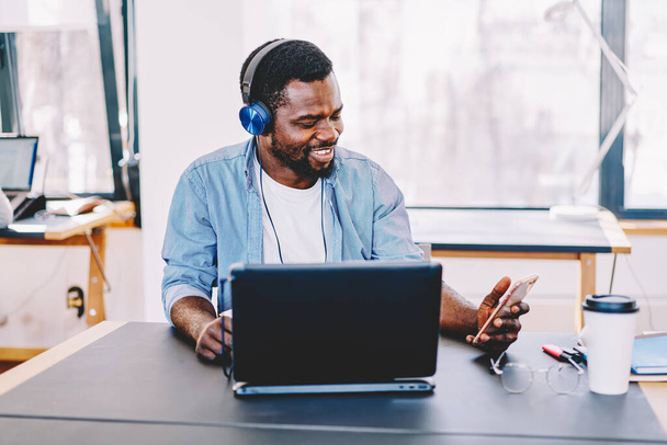 Happy successful black man in headphones using cellphone while working and sitting with laptop at table in light modern office - 写真・画像