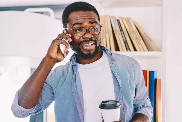 Cheerful African American male employee in glasses talking on cellphone and holding takeaway cup while standing in light modern office - Photo, Image