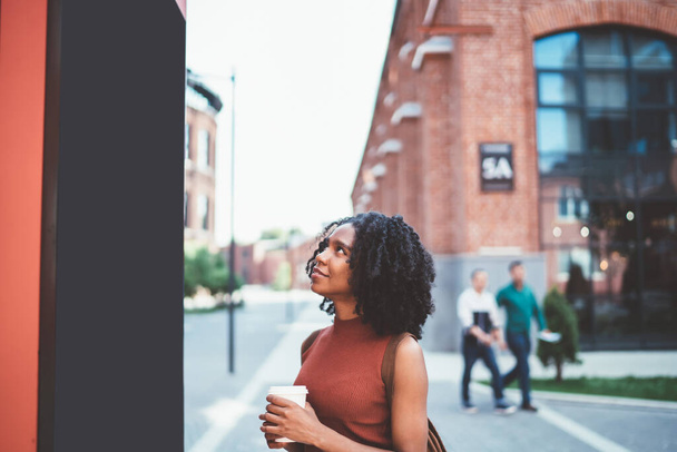 Viaggiatore afro-americano sognatore con acconciatura afro in abiti casual guardando cartellone prendendo il caffè esplorare la città passeggiando per strada - Foto, immagini