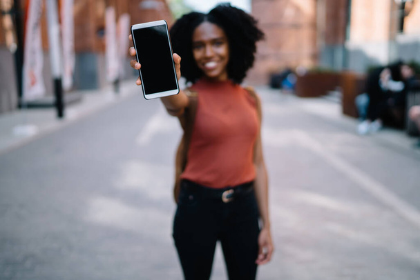 Glad black curly haired female in casual wear showing display of modern phone spending time strolling in street of city - Photo, Image