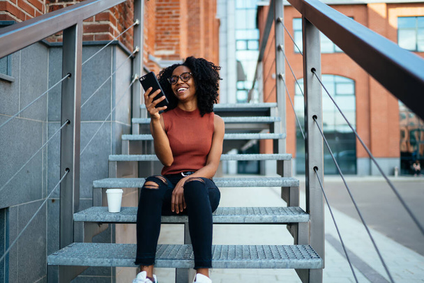 Young spectacled African American female looking at mobile phone screen and cheerfully smiling while having rest on stair in city with cup of coffee - 写真・画像