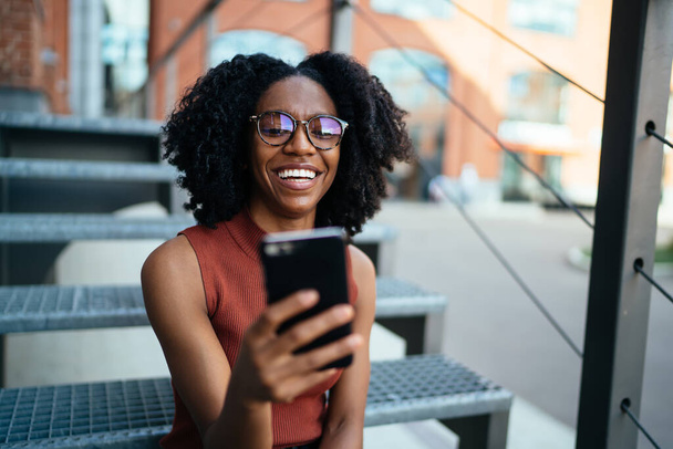 Happy African American laughing lady in casual clothes and glasses with Afro hairstyle watching mobile sitting on stairs spending time in urban street - Zdjęcie, obraz