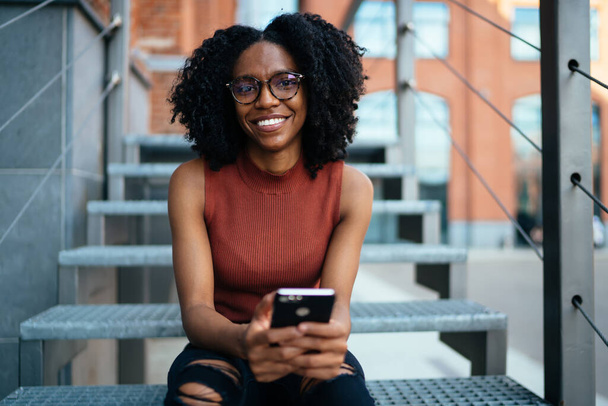 Bright African American smiling female in casual clothes and glasses with Afro hairstyle using mobile messaging with friend sitting on stairs in street of modern city looking at camera - Fotoğraf, Görsel