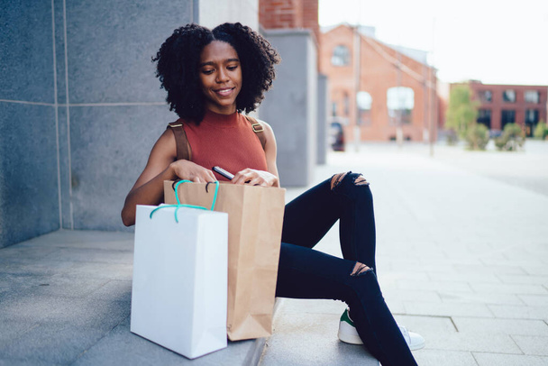 Cheerful content white toothed African American curly haired woman sitting on stairs with paper bags in sleeveless casual shirt in street in summer - Foto, Imagem