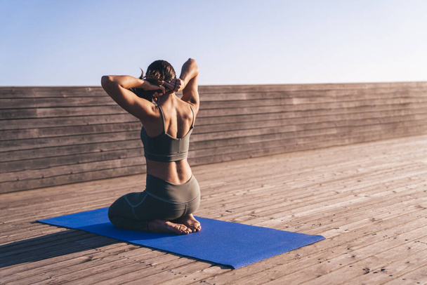 Back view of peaceful shapely athletic woman in modern cozy sportswear practicing yoga on blue comfy mat in bright daylight - Fotoğraf, Görsel