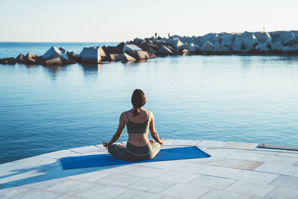 Back view of lady in yoga clothing sitting on blue mat in lotus position on stone pier in morning near calm water - Photo, Image