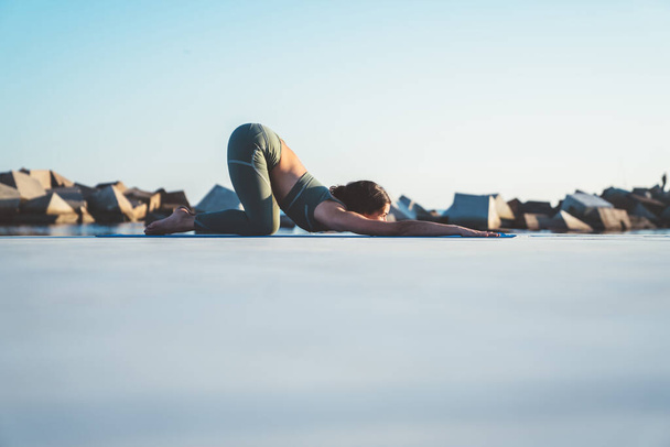 Vue latérale de la femme mince en vêtements de sport faisant du yoga en position demi-tortue sur le tapis avec mer floue et pierres sur le fond - Photo, image