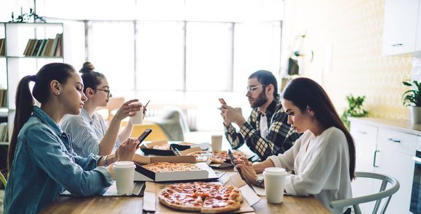 Side view of serious friends browsing mobile phones during meeting sitting at table with fresh tasty pizza and coffee in canteen - 写真・画像