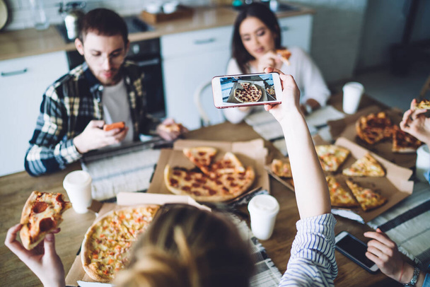High angle selective focus of anonymous woman taking photo of pizza while sitting at table with friends and eating during party - Foto, Bild