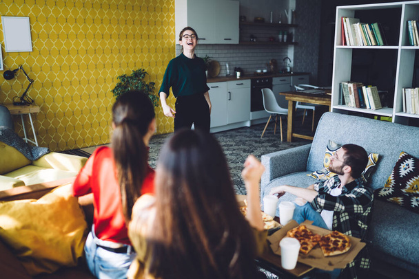 Positive friends in casual wear eating appetizing pizza and looking at laughing friend while relaxing on soft comfy sofa in modern apartment - Foto, imagen
